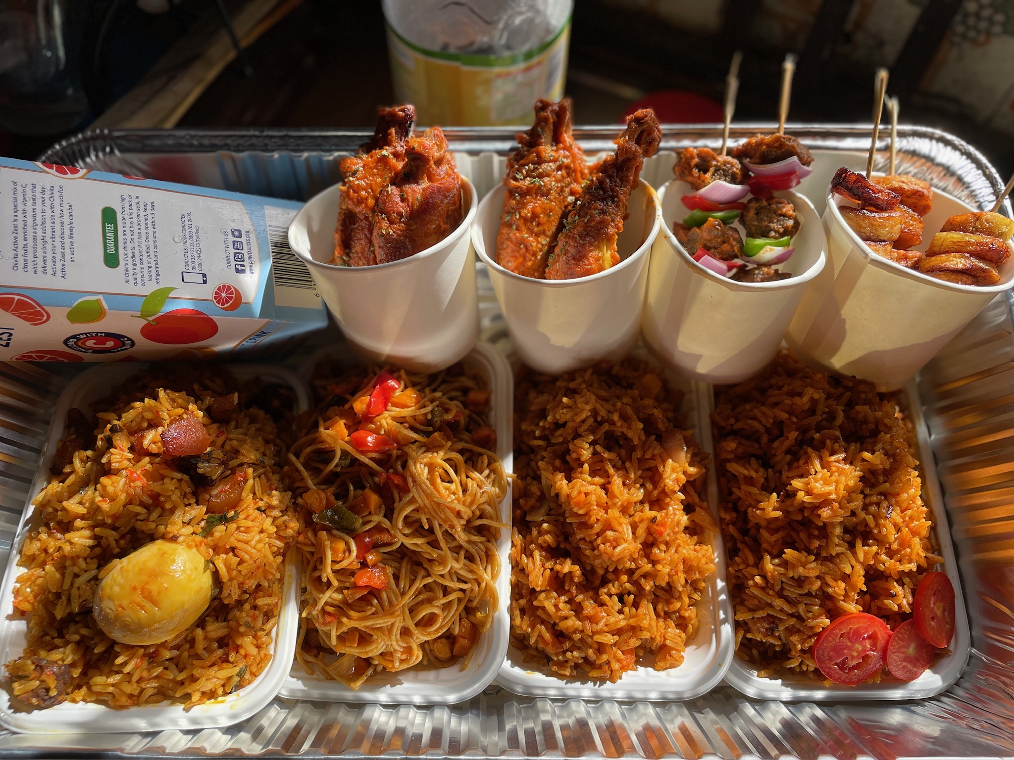 A catered spread of jollof, fried rice, spaghetti and proteins in trays